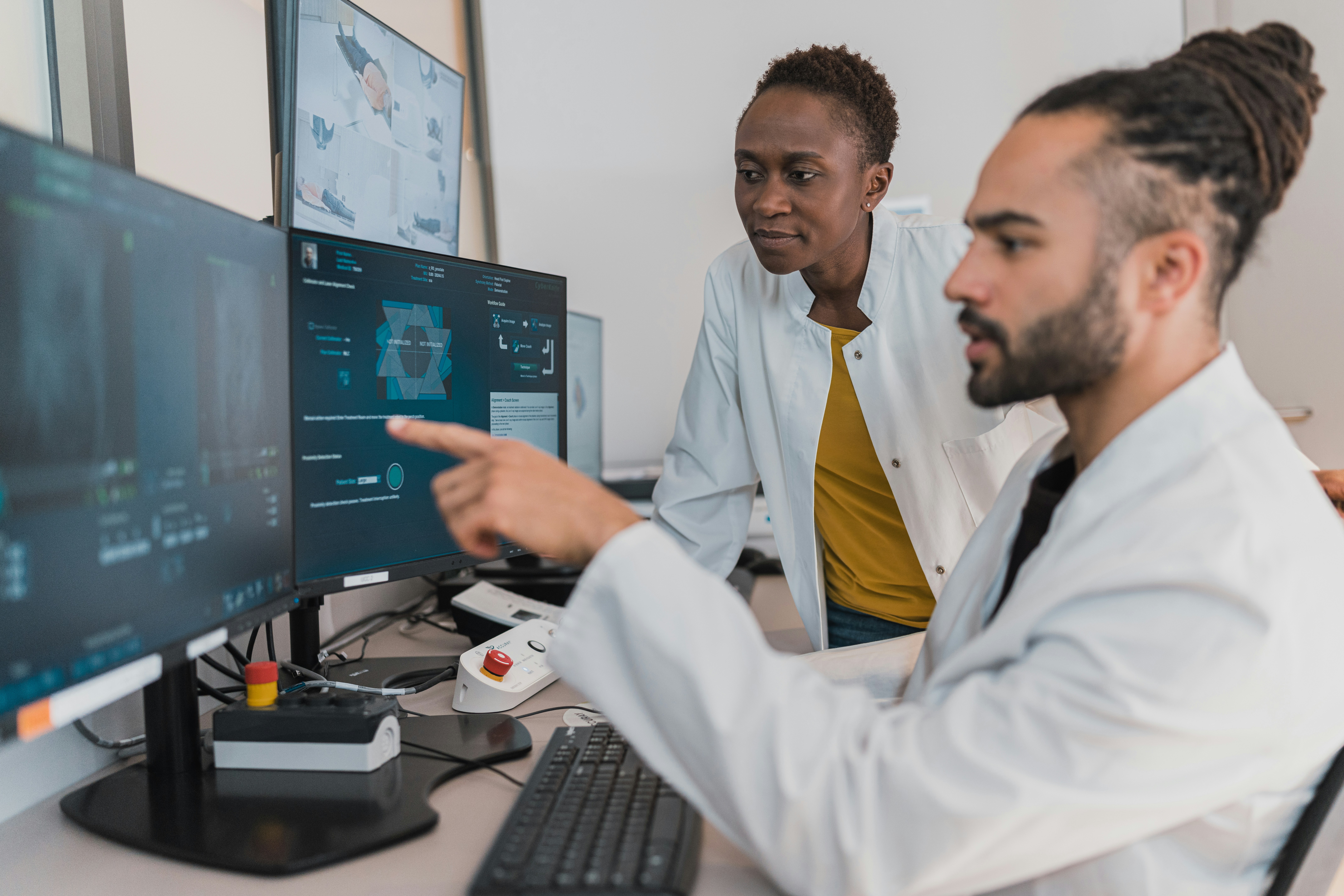 Two medical researchers in white lab coats examine data on multiple computer monitors, with one pointing to a digital interface on the screen.